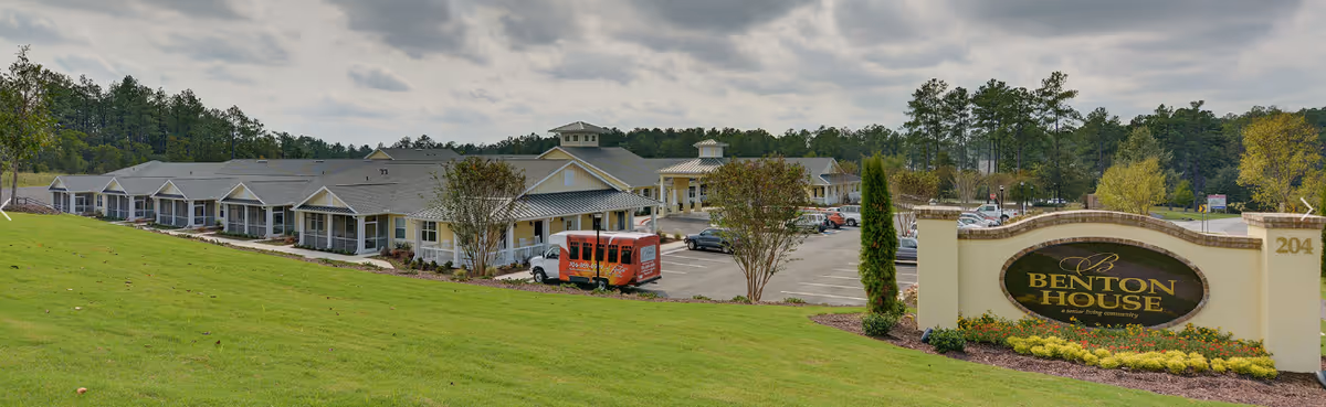 Wide exterior view of Benton House senior living community showing a large single-story building with multiple entrances, a parking lot with cars, a red shuttle van, and a landscaped grassy area with trees under a cloudy sky. A large sign in the foreground reads 'Benton House'.