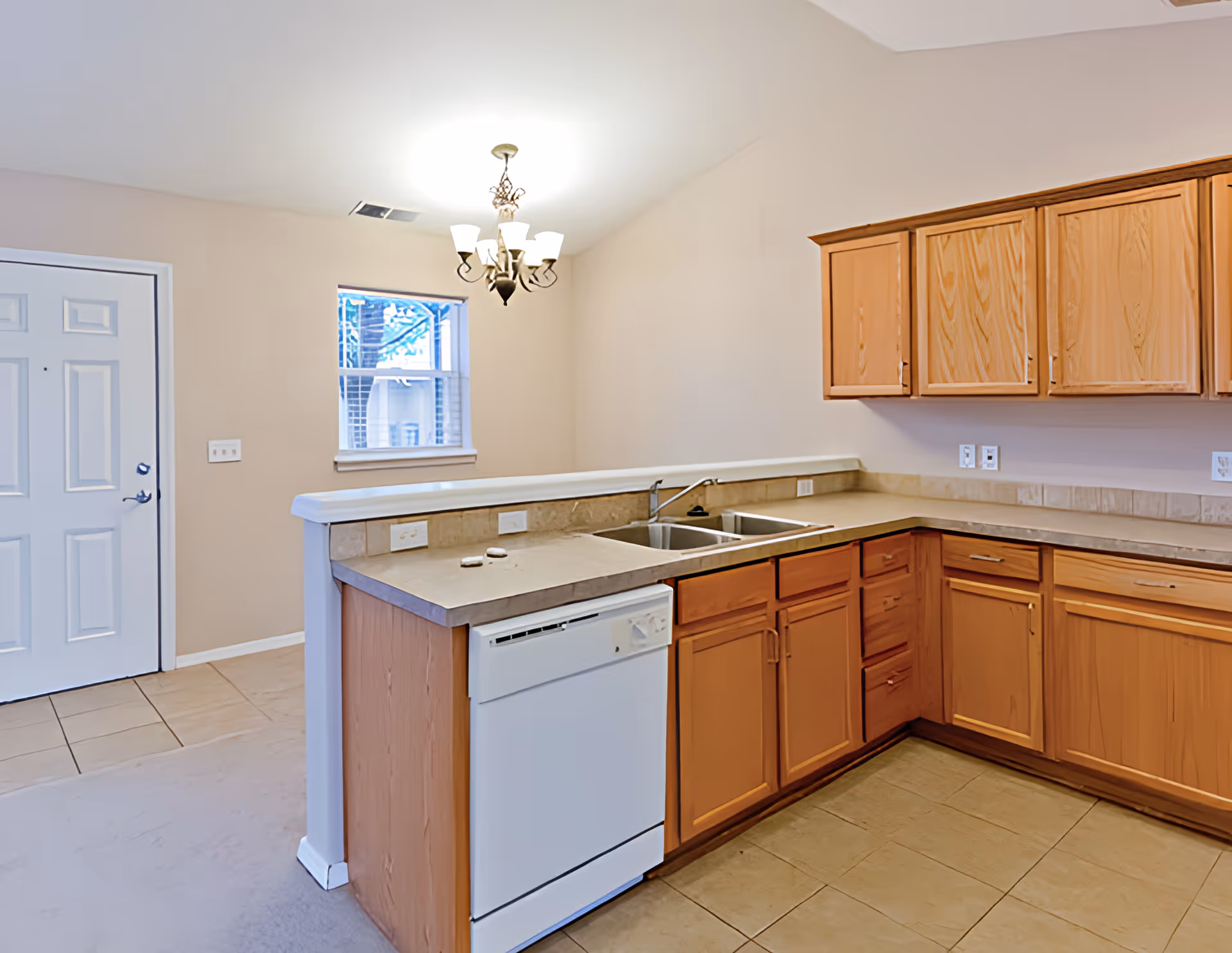 Open kitchen with light wood cabinets, L-shaped countertops, a double sink and dishwasher, and an entry door with a small window and chandelier.