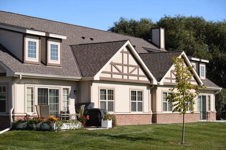 Exterior front of a single-story residential-style building with gabled roof, windows, patio furniture, flowers, and a small lawn with a young tree.