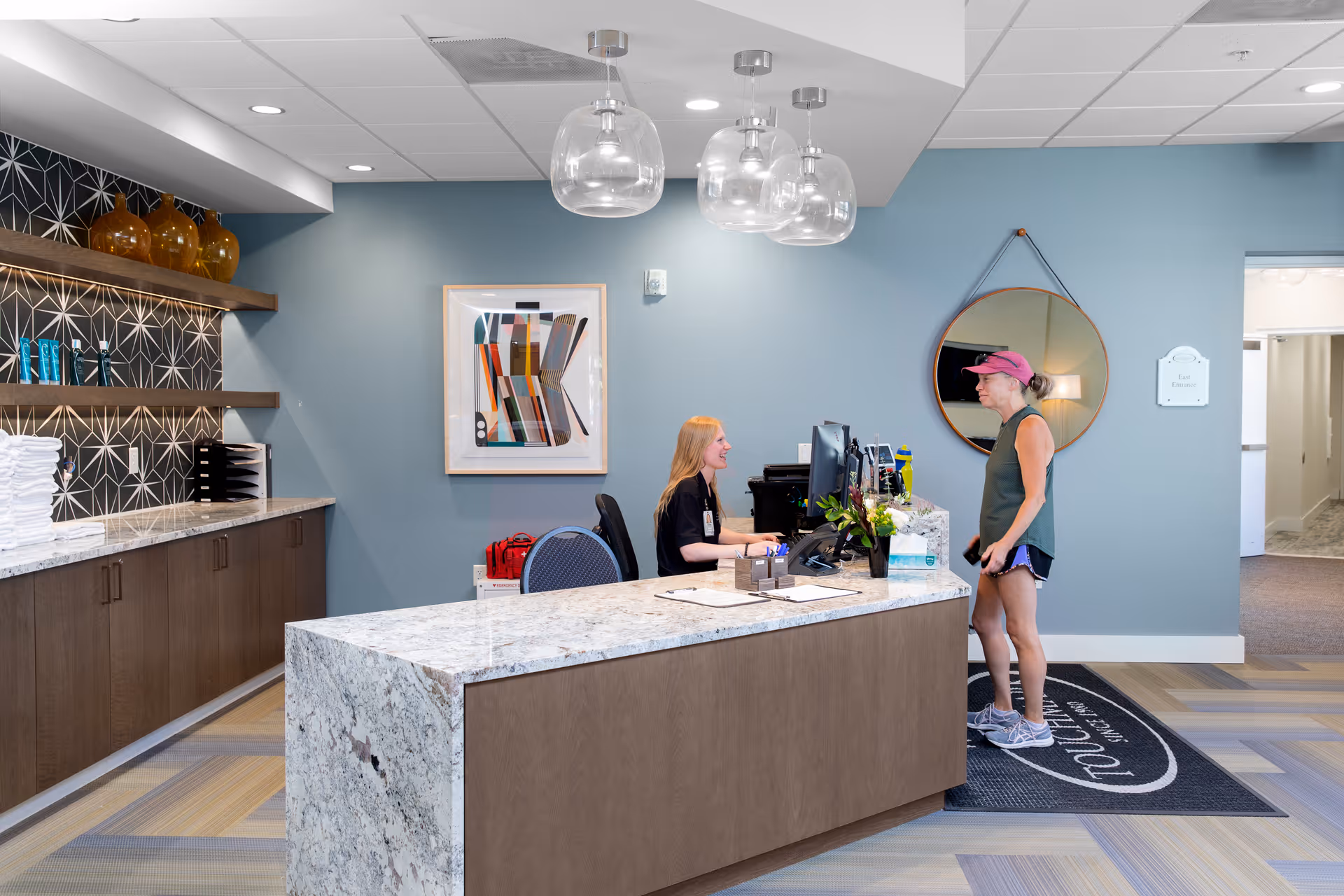 Reception area in a senior living facility with a woman sitting behind a marble reception desk talking to a visitor wearing athletic clothes and a pink cap. The room has blue walls, modern pendant lights, a round mirror, abstract artwork, and shelves with decorative items and folded towels.
