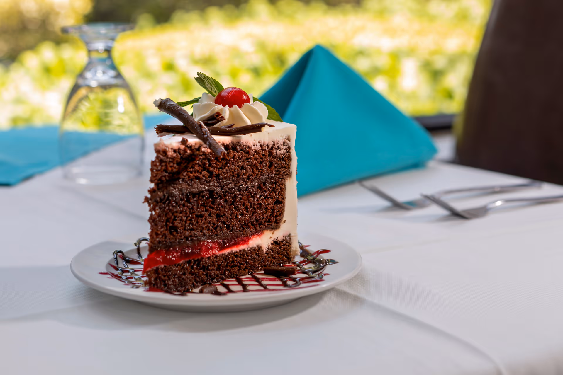 A slice of chocolate cake with white frosting, a cherry on top, and chocolate decorations on a white plate with chocolate drizzle, placed on a white tablecloth with a folded teal napkin, a glass, and silverware in the background.