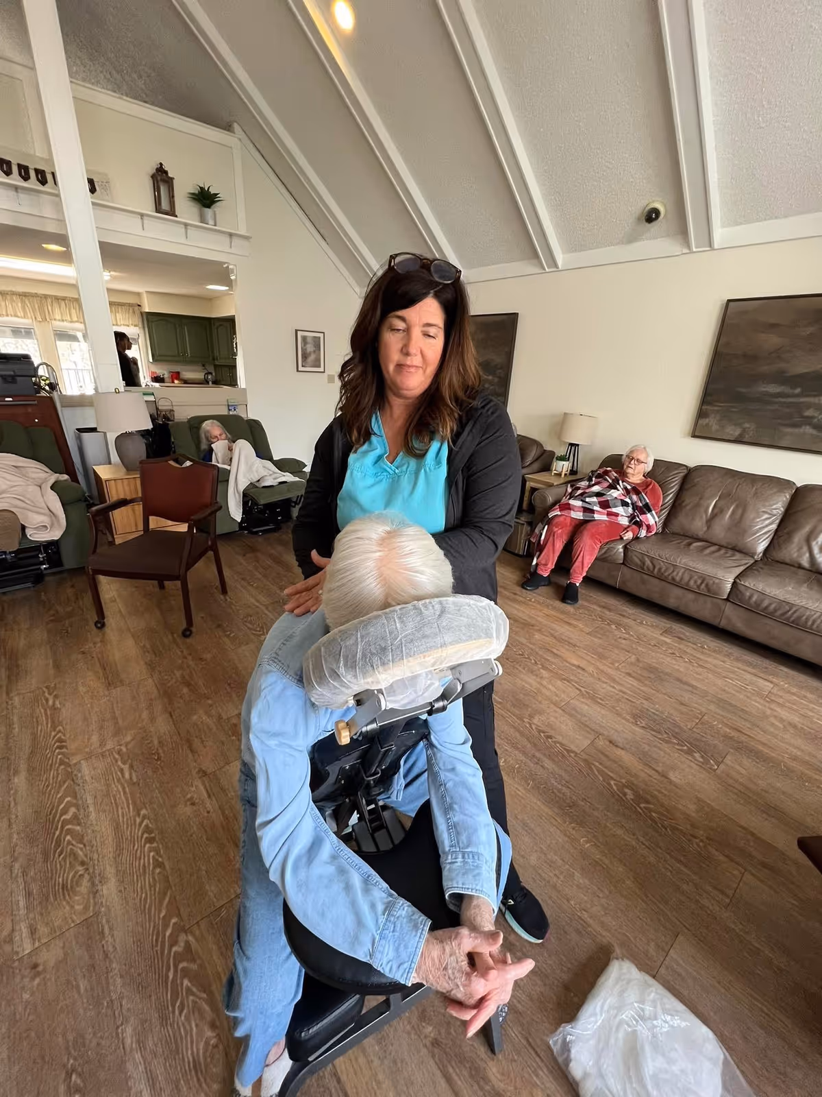 A woman in a teal shirt and black jacket is giving a seated massage to an elderly person with white hair in a living room setting. In the background, two elderly individuals are seated on chairs and a brown leather couch, one covered with a blanket. The room has wooden flooring, high vaulted ceilings, and neutral-colored walls with framed artwork.