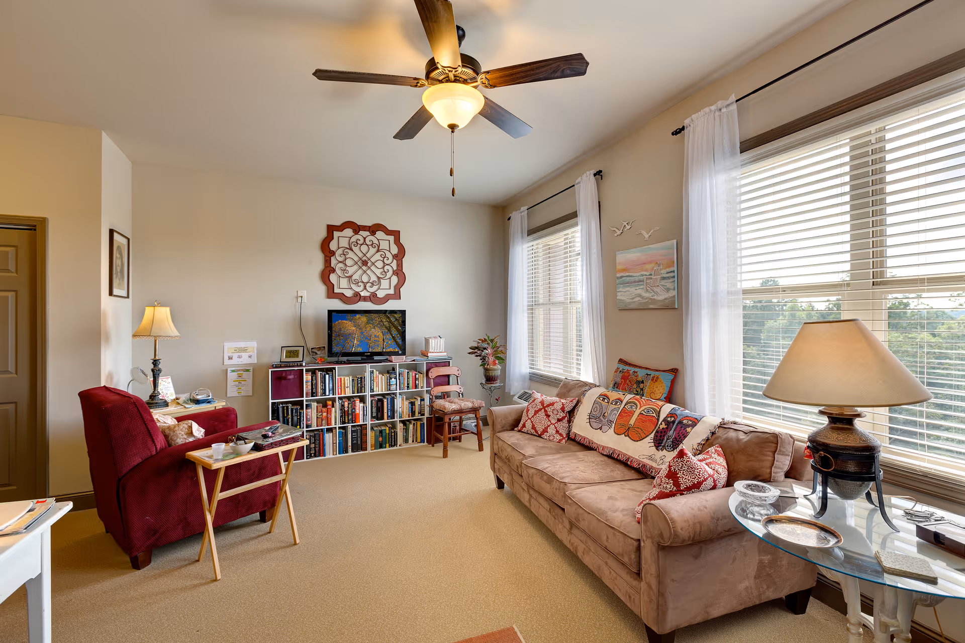 A cozy living room with a beige couch adorned with colorful owl-themed pillows, a red armchair with a small wooden tray table, a bookshelf filled with books and a TV on top, two large windows with white curtains letting in natural light, a ceiling fan with a light fixture, and two table lamps providing additional lighting.