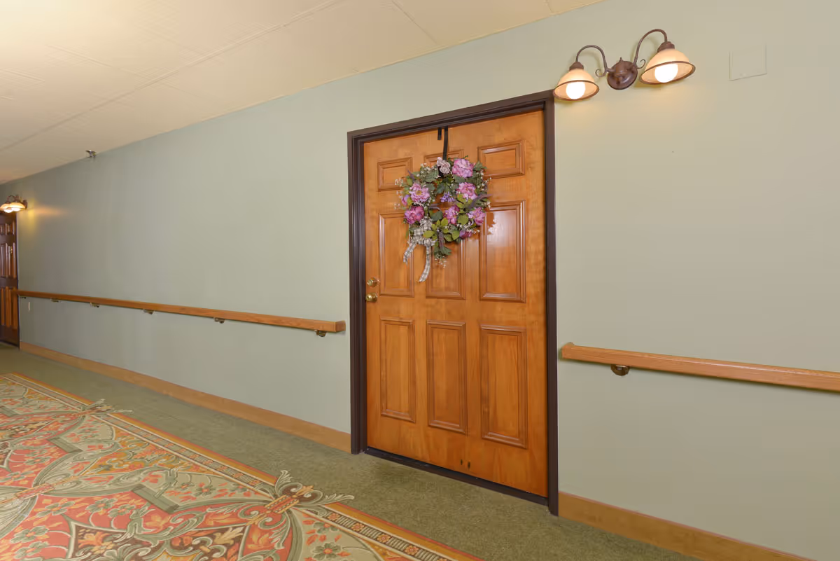 A hallway in a senior living facility with a wooden door decorated with a floral wreath. The walls are painted light green with wooden handrails on both sides. There are two wall-mounted light fixtures above the door and another light fixture further down the hallway. The floor is carpeted with a patterned design near the door.