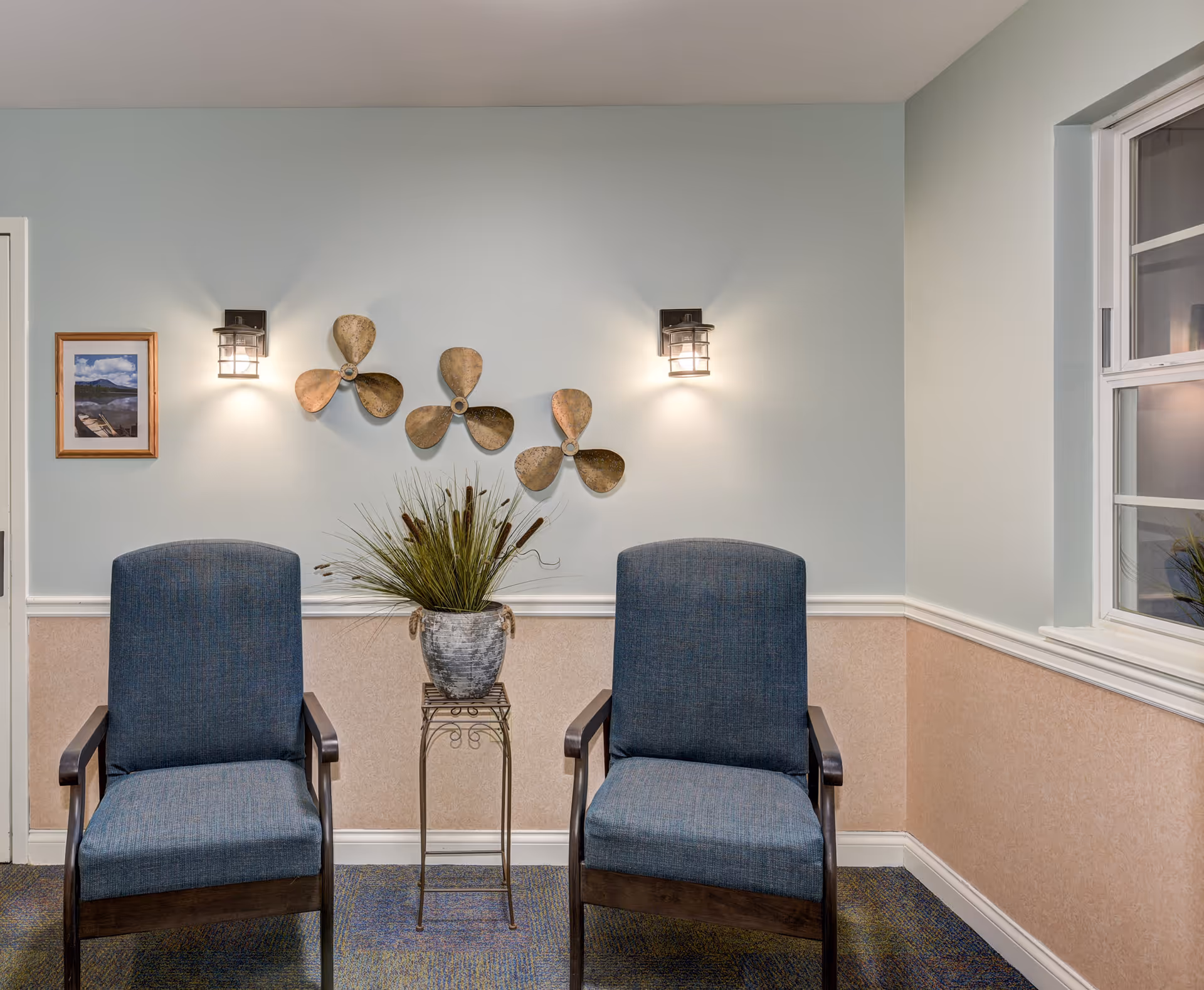 A small seating area with two blue upholstered chairs with wooden arms, separated by a small metal stand holding a decorative plant. The wall behind is painted light blue on the upper half and beige on the lower half, with white trim in between. Three decorative metal propellers are mounted on the wall between two wall-mounted light fixtures. A framed picture hangs on the left side of the wall, and a window is visible on the right side.