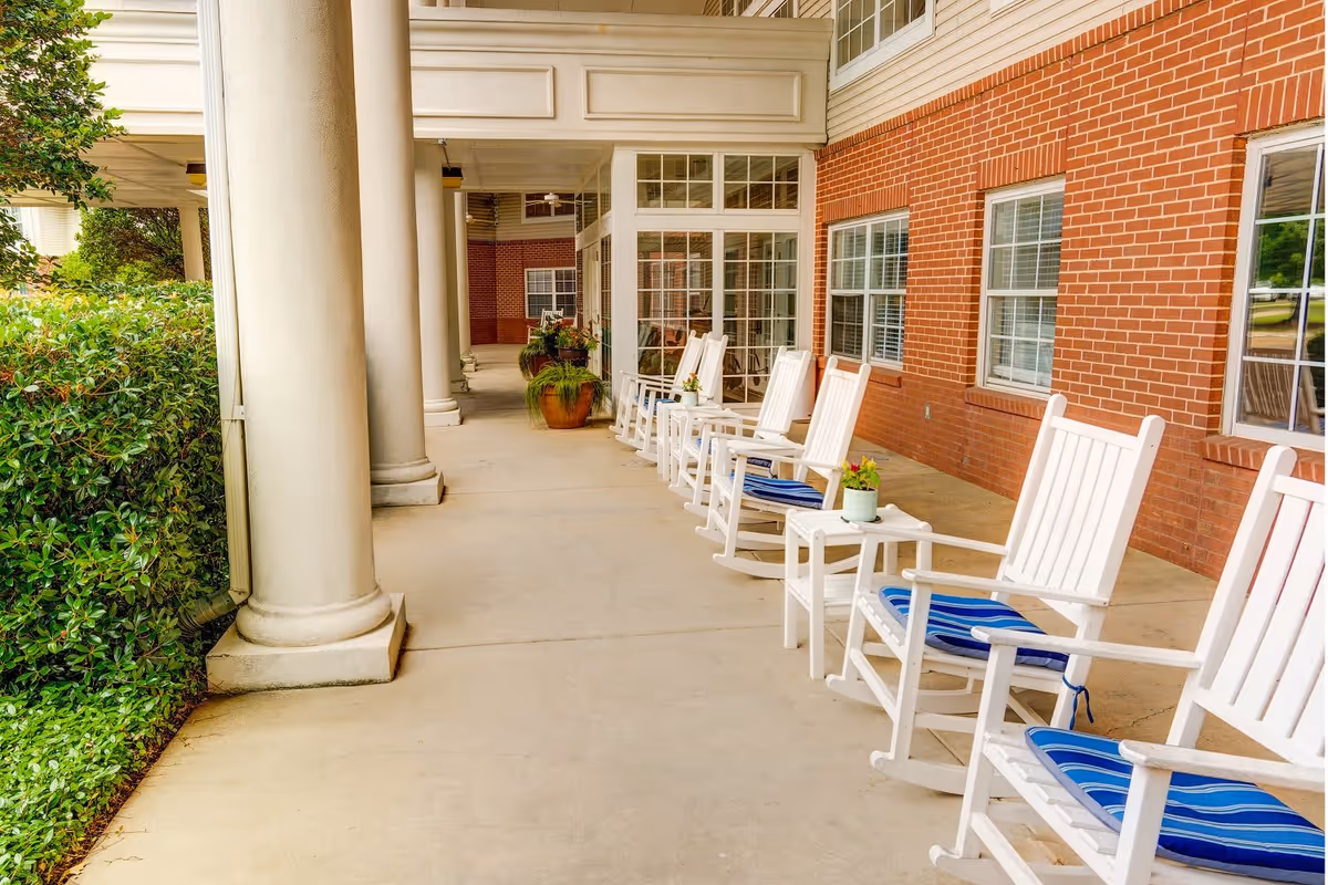 A covered outdoor patio area with white rocking chairs lined up along a brick wall. Each chair has a blue cushion, and small tables with potted plants are placed between some of the chairs. Large white columns support the roof, and greenery is visible on the left side.
