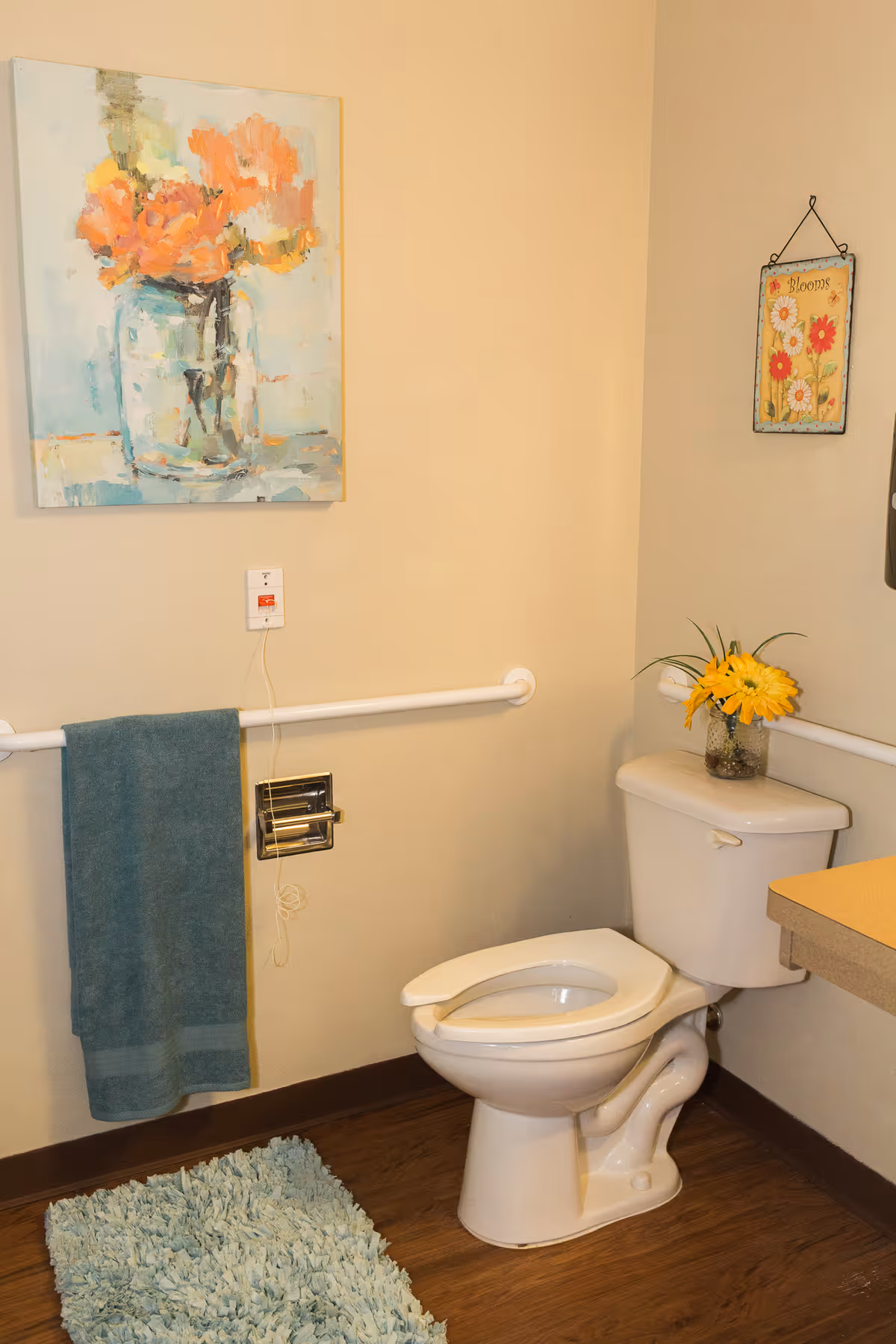 A clean bathroom with a white toilet, a blue towel hanging on a grab bar, a blue shaggy rug on the wooden floor, and two floral decorations on the walls and toilet tank.