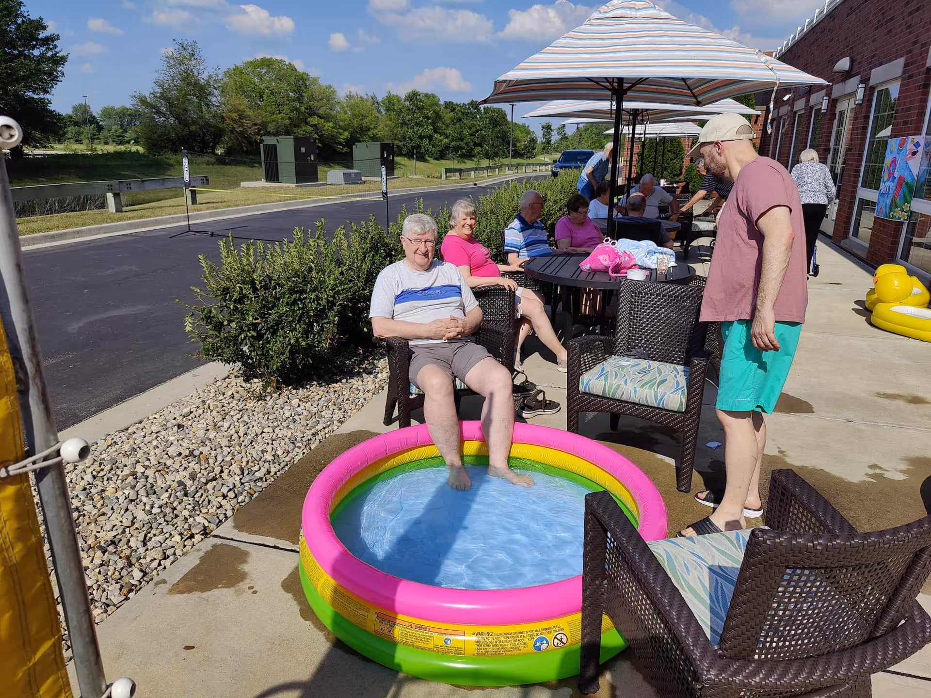 A group of elderly people and a younger man are enjoying a sunny day outside a brick building. One elderly man is sitting with his feet in a small inflatable kiddie pool filled with water. Several people are seated around tables under large patio umbrellas, and there are chairs arranged on the concrete patio. The background shows a parking lot, greenery, and a clear blue sky with some clouds.