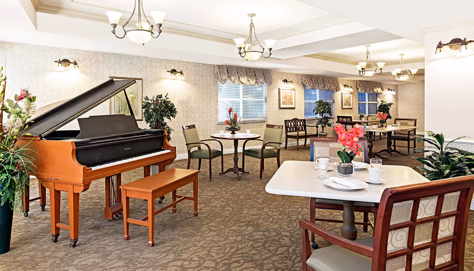 A spacious senior living common area with multiple tables and chairs arranged for socializing or dining. A grand piano with a matching bench is positioned on the left side, surrounded by plants and decorative flowers. The room is well-lit with ceiling chandeliers and wall sconces, and windows with valances let in natural light.