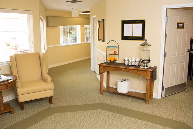 Bright interior common area with a beige wingback chair, side table, and a wooden console table holding a beverage dispenser and cups.