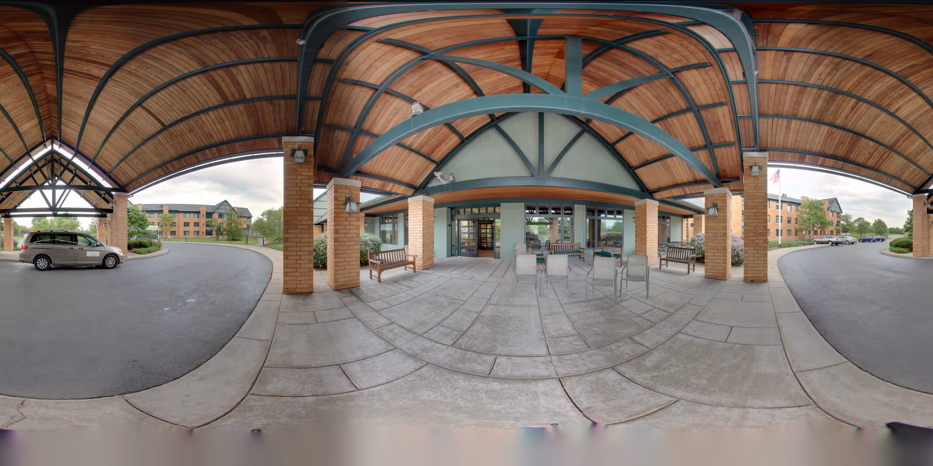 Covered entrance area of The Summit at Brighton Independent Living facility with a wooden arched ceiling supported by brick pillars. There are benches and chairs arranged under the covered area. A driveway curves around the entrance with a vehicle parked on the left side. The building exterior is visible in the background with multiple windows and greenery around.