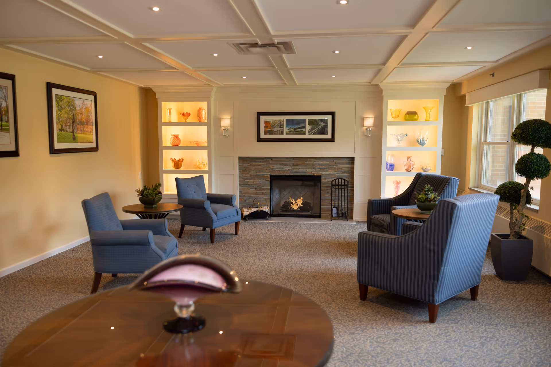 A cozy living room with four blue upholstered armchairs arranged around small wooden tables. The room features a fireplace with a stone surround and built-in shelves on either side displaying decorative glass items. There are framed pictures on the walls and a large window letting in natural light. A potted topiary plant is placed near the window.
