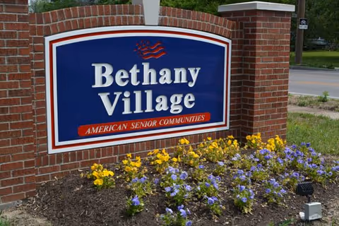A brick sign for Bethany Village, an American senior community, surrounded by a flower bed with yellow and purple flowers, located near a road.