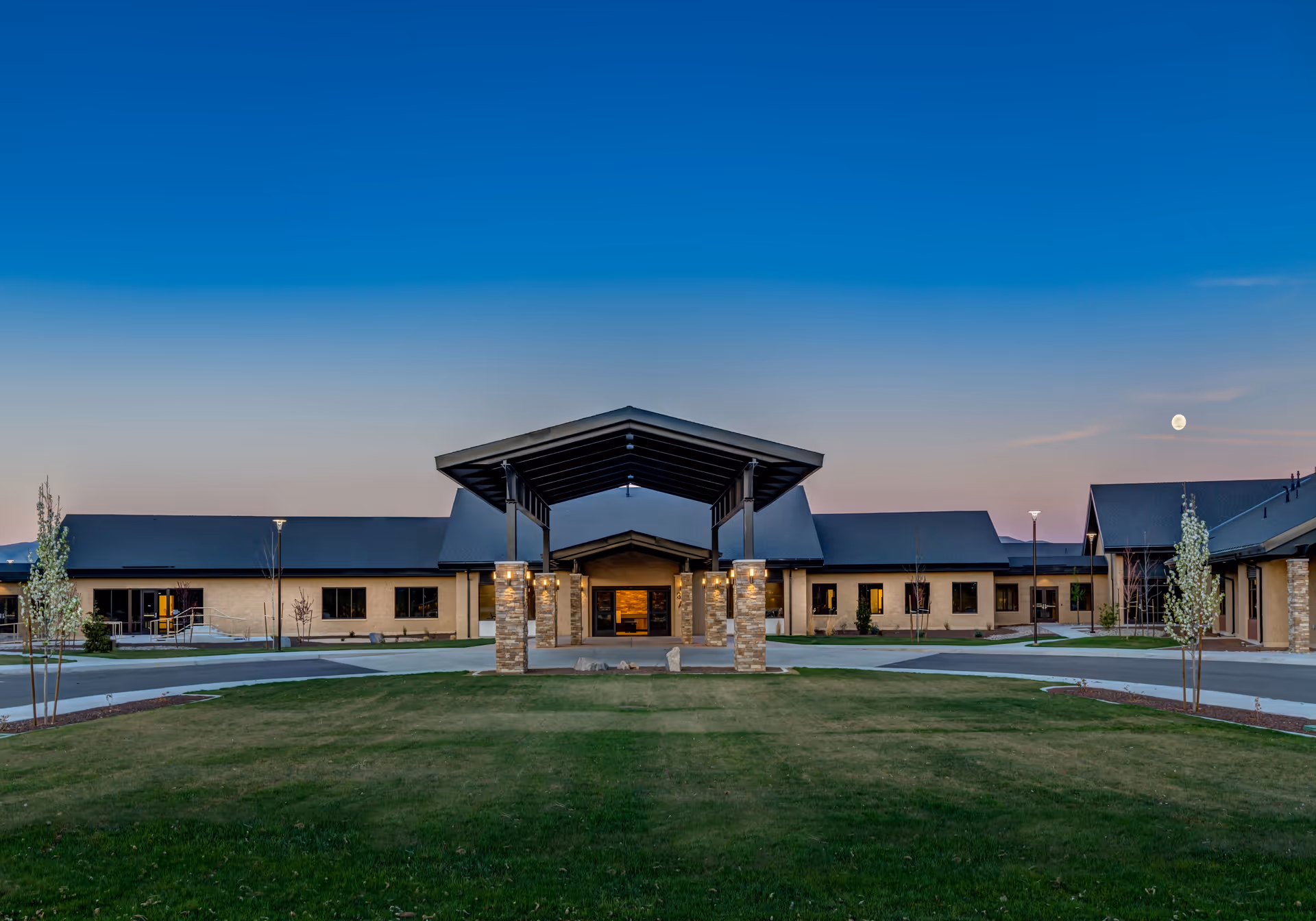 Exterior view of the Northern Nevada State Veterans Home building at dusk, showing a large covered entrance with stone pillars, a well-maintained lawn in the foreground, and a clear sky with the moon visible.