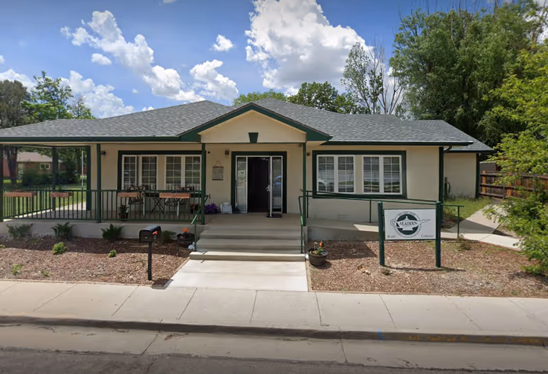 Front exterior view of a single-story assisted living facility building with a covered porch, steps leading to the entrance, and a sign that reads Aladdin Assisted Living. The building is surrounded by some landscaping and trees under a partly cloudy sky.