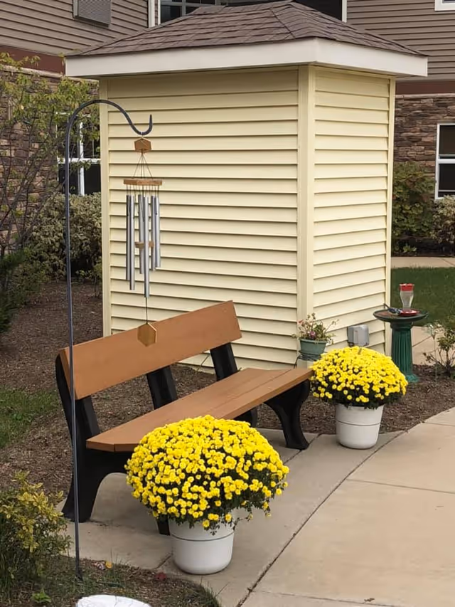 Outdoor seating area with a brown bench and two large pots of yellow flowers placed on either side. A wind chime hangs from a metal stand next to the bench. The background shows a small beige shed with siding and part of a building with stone and siding exterior.