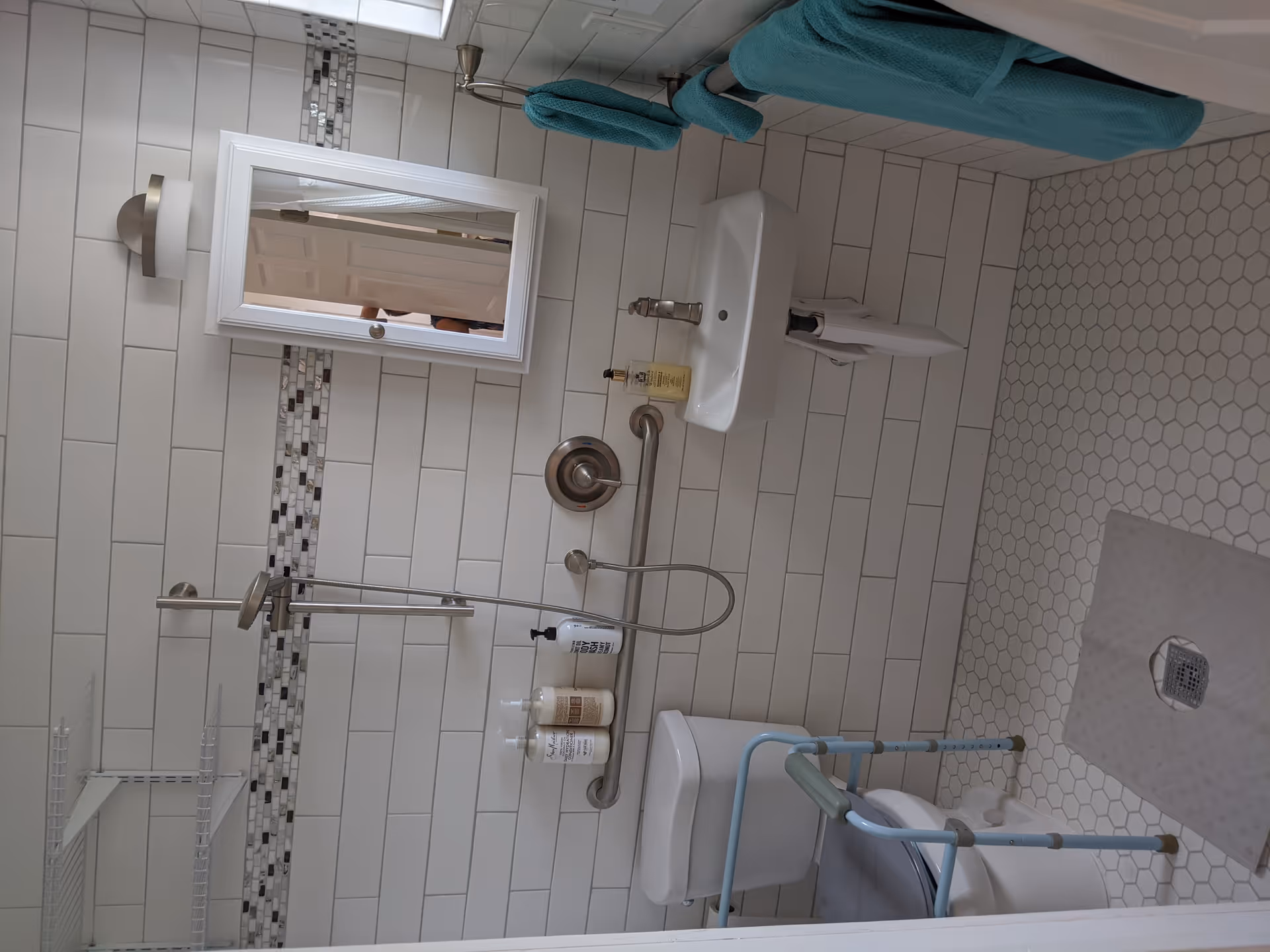 A clean, accessible bathroom with white subway tiles on the walls and hexagonal white floor tiles. The bathroom features a wall-mounted sink with a soap dispenser, a toilet with a blue support frame, a shower area with a handheld showerhead, and a grab bar. There is a white framed mirror above the sink and teal towels hanging on a towel rack.