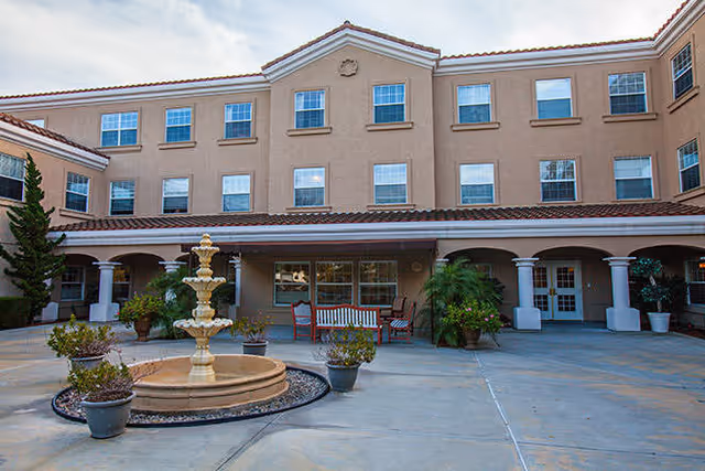 Exterior view of a three-story building with beige walls and a red-tiled roof, featuring multiple windows. In front of the building is a courtyard with a multi-tiered fountain surrounded by potted plants and outdoor seating including a bench and chairs. The sky is partly cloudy.