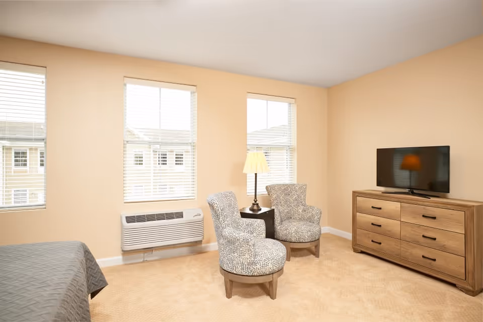 A cozy room with beige walls and carpet featuring two patterned armchairs around a small table with a lamp, a wooden dresser with a flat-screen TV on top, and three windows with white blinds letting in natural light.