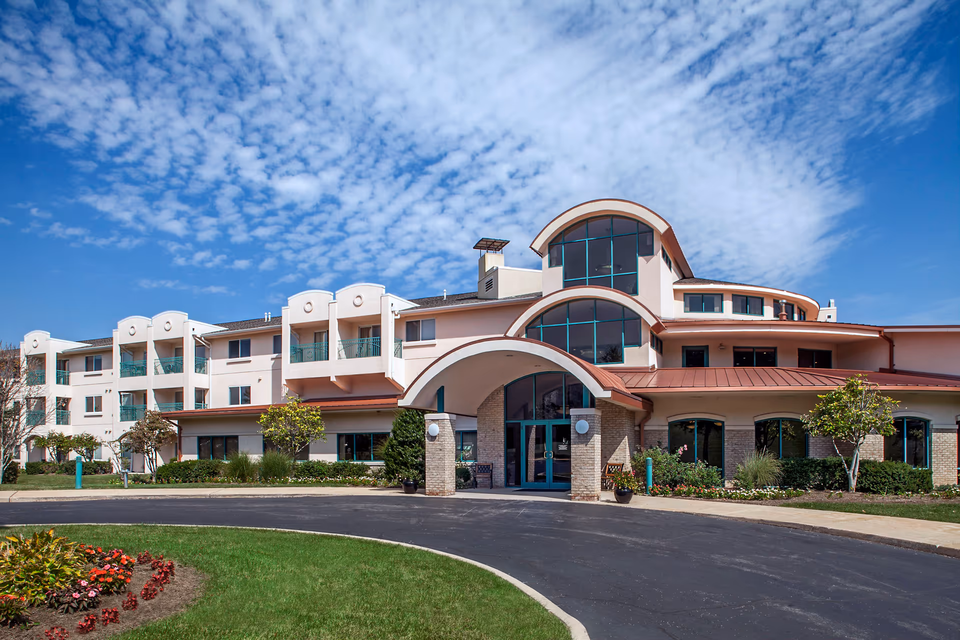 Front entrance of a multi-story senior living building with arched windows, balconies, landscaping and a curved driveway under a blue sky.