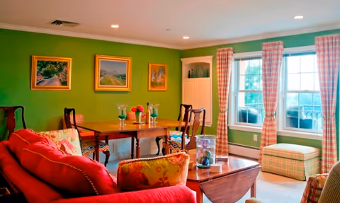 A cozy living room with a red sofa adorned with floral cushions, a wooden coffee table with decorative items, a wooden dining table with chairs, green walls with three framed paintings, and large windows with red and white checkered curtains letting in natural light.