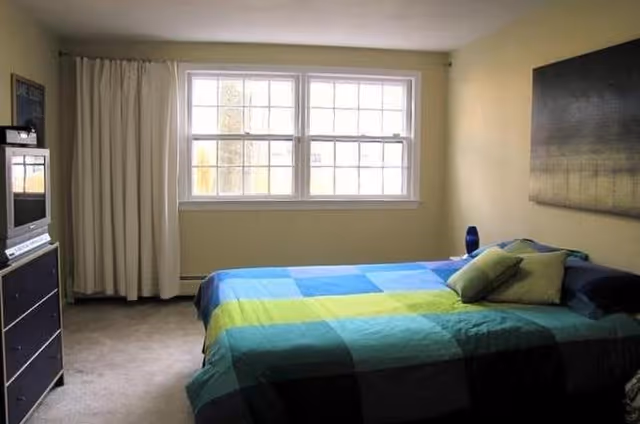 A bedroom with a large window covered by white curtains, a bed with a colorful blue and green checkered bedspread and pillows, a dark dresser with a TV on top, and a large piece of artwork hanging on the wall.