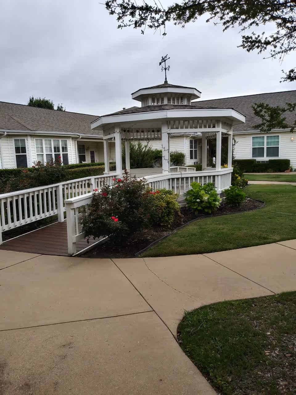 Outdoor view of a white gazebo with a weather vane on top, surrounded by bushes and flowers, located in the courtyard of a senior living facility with beige buildings and windows in the background under a cloudy sky.