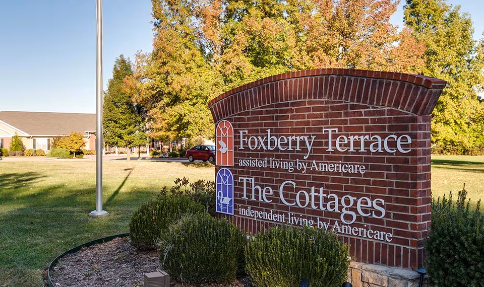 Brick entrance sign reading 'Foxberry Terrace' and 'The Cottages' on a grassy lawn with trees and a building in the background.
