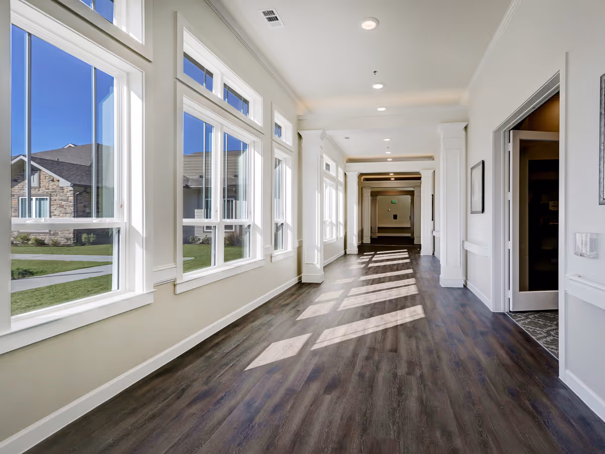 Bright hallway with large windows on the left side letting in natural light, white walls, and dark wood flooring. The hallway leads to a distant door with multiple doorways along the right side.