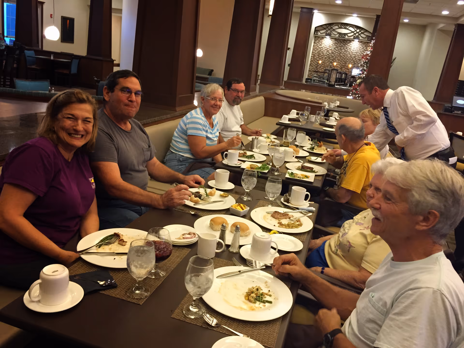 A group of seniors and a staff member seated around a long dining table enjoying a meal in a well-lit dining room.