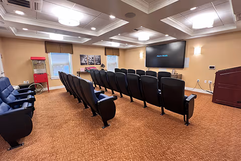 A small theater-style room with two rows of black cushioned chairs facing a large flat screen TV mounted on a beige wall. The room has a popcorn machine in the corner, two windows with brown valances, and warm lighting from ceiling fixtures and wall sconces. The carpet is a reddish-brown color, and there is a wooden podium on the right side of the room.