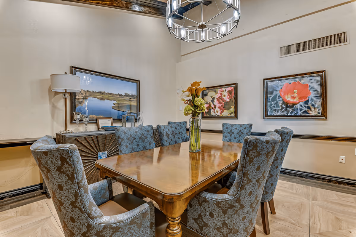 A dining room with a polished wooden table surrounded by eight upholstered chairs with a blue and brown pattern. A vase with flowers is placed in the center of the table. The walls are decorated with framed pictures of flowers and a landscape. A modern chandelier hangs from the ceiling, and a sideboard with a lamp and decorative items is against one wall.
