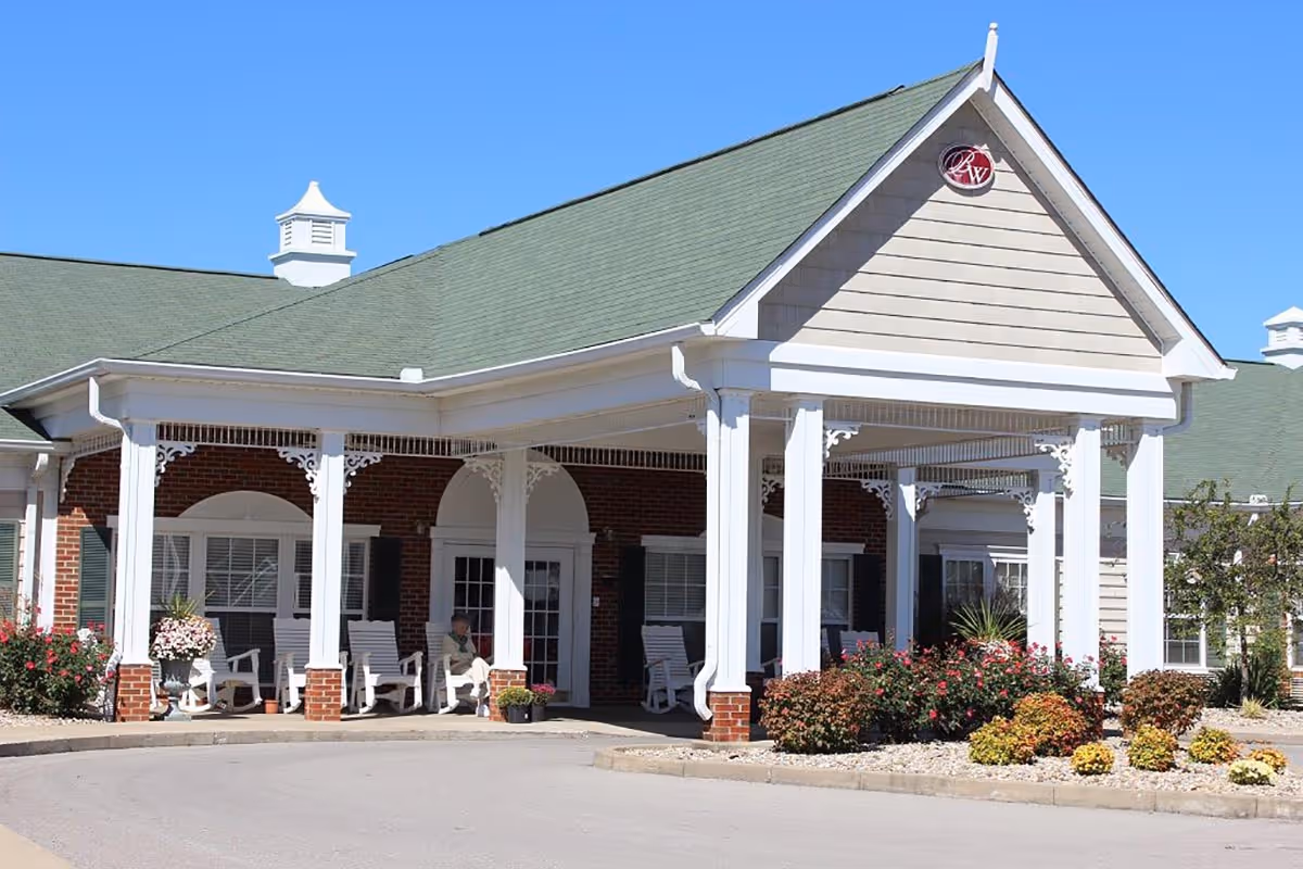 Exterior view of Bluegrass Way Senior Living facility showing a covered entrance with white columns and rocking chairs. There are bushes and flowers planted around the entrance, and a person is sitting on one of the rocking chairs. The building has a green roof and brick walls with white trim.