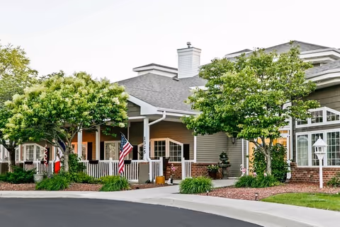 Exterior view of a residential building with a porch, American flag, and well-maintained landscaping including trees and shrubs, under a clear sky.