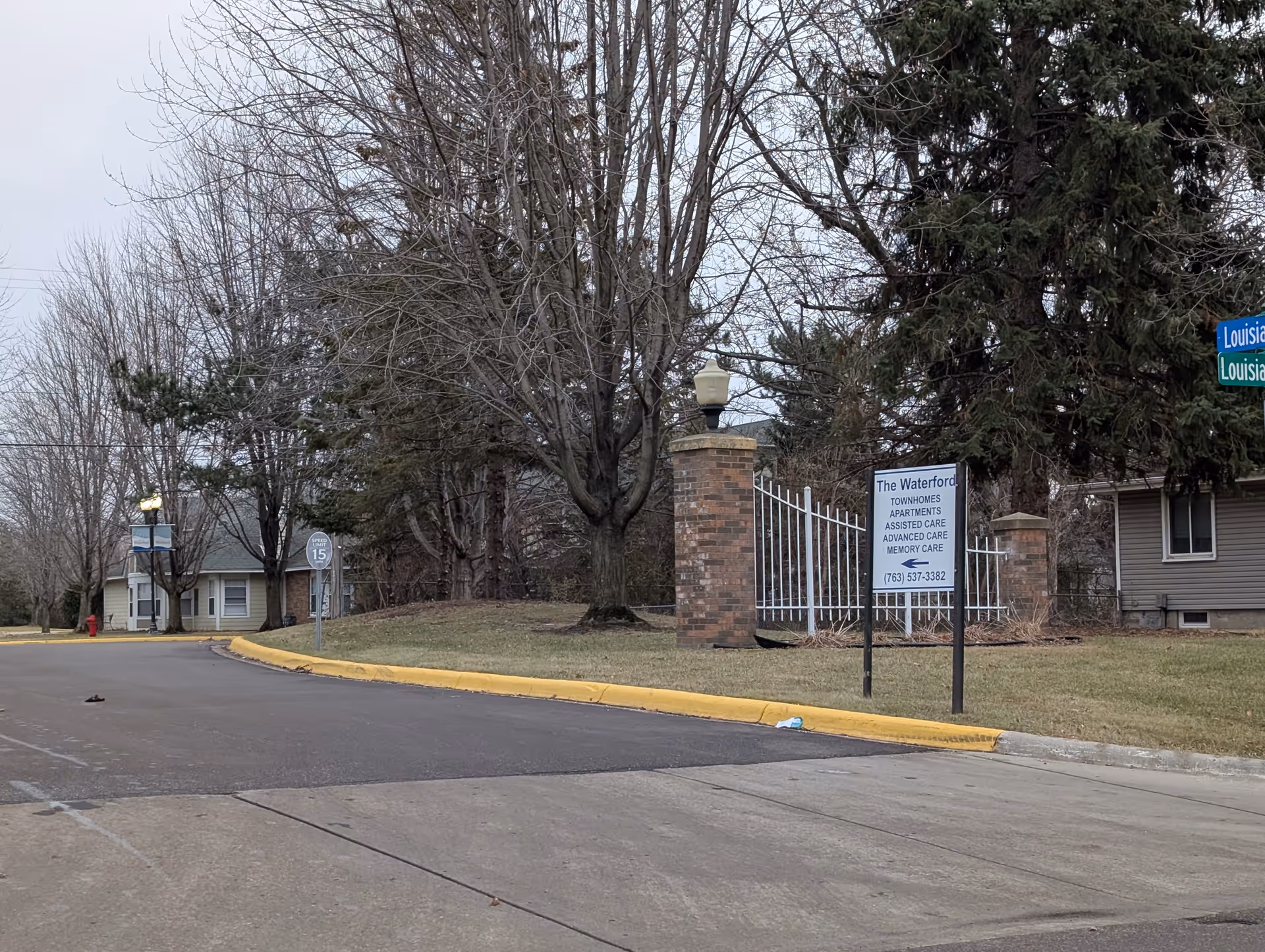 Street view of the entrance to The Waterford senior living facility, showing a brick pillar with a lamp, a white metal gate, a sign listing townhomes, apartments, assisted care, advanced care, and memory care, and nearby houses and leafless trees on a cloudy day.