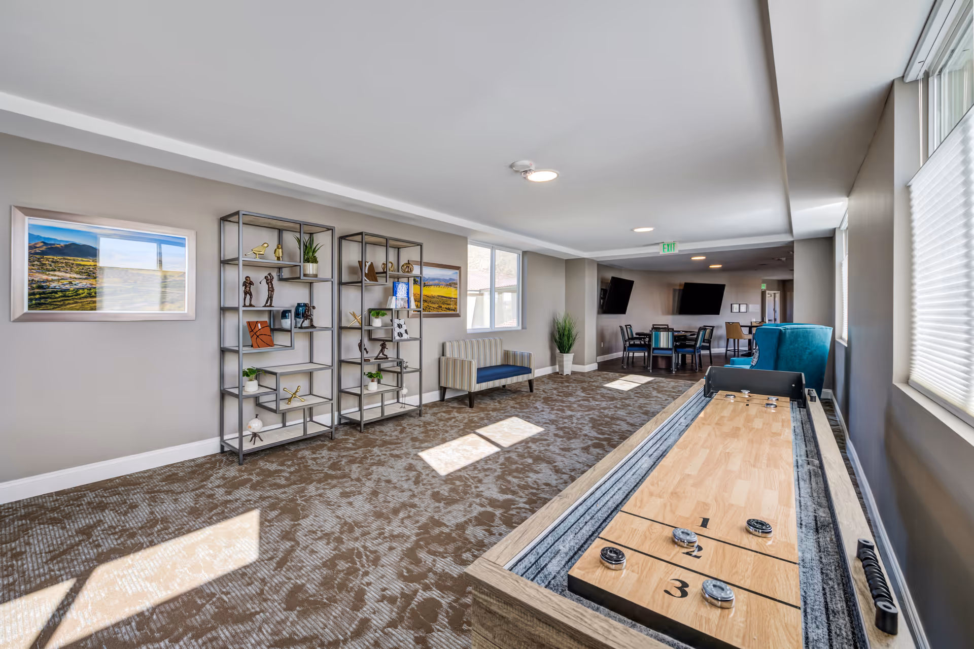 A bright and spacious common area in a senior living facility featuring a shuffleboard table in the foreground, two modern metal shelving units with decorative items, a striped loveseat, and a seating area with tables and chairs near two wall-mounted televisions. Large windows allow natural light to fill the room, which has beige walls and patterned carpet.