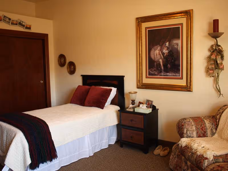 A cozy bedroom with a single bed covered in a white quilt and two red pillows. Next to the bed is a dark wooden nightstand with two drawers, holding a lamp, a tissue box, and some small items. On the wall above the nightstand is a large framed painting. To the right is a patterned armchair with a cream-colored throw blanket. The room has beige walls and carpeted floor, with a closed wooden door and two small round wall decorations on the left.