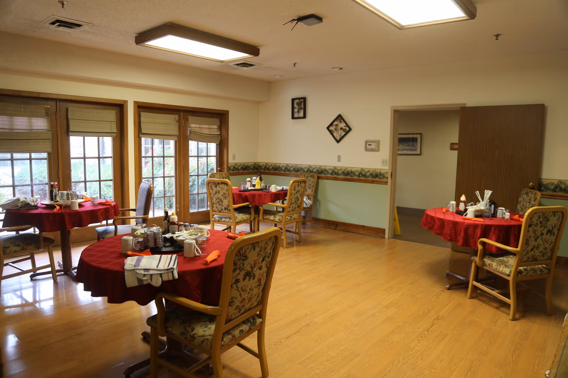 A dining room with four round tables covered with red tablecloths, each surrounded by floral upholstered chairs. The tables have various condiments, cups, and napkins on them. Large windows with blinds allow natural light to enter, and there are framed pictures on the walls. The floor is wooden, and there is an open door leading to another room.