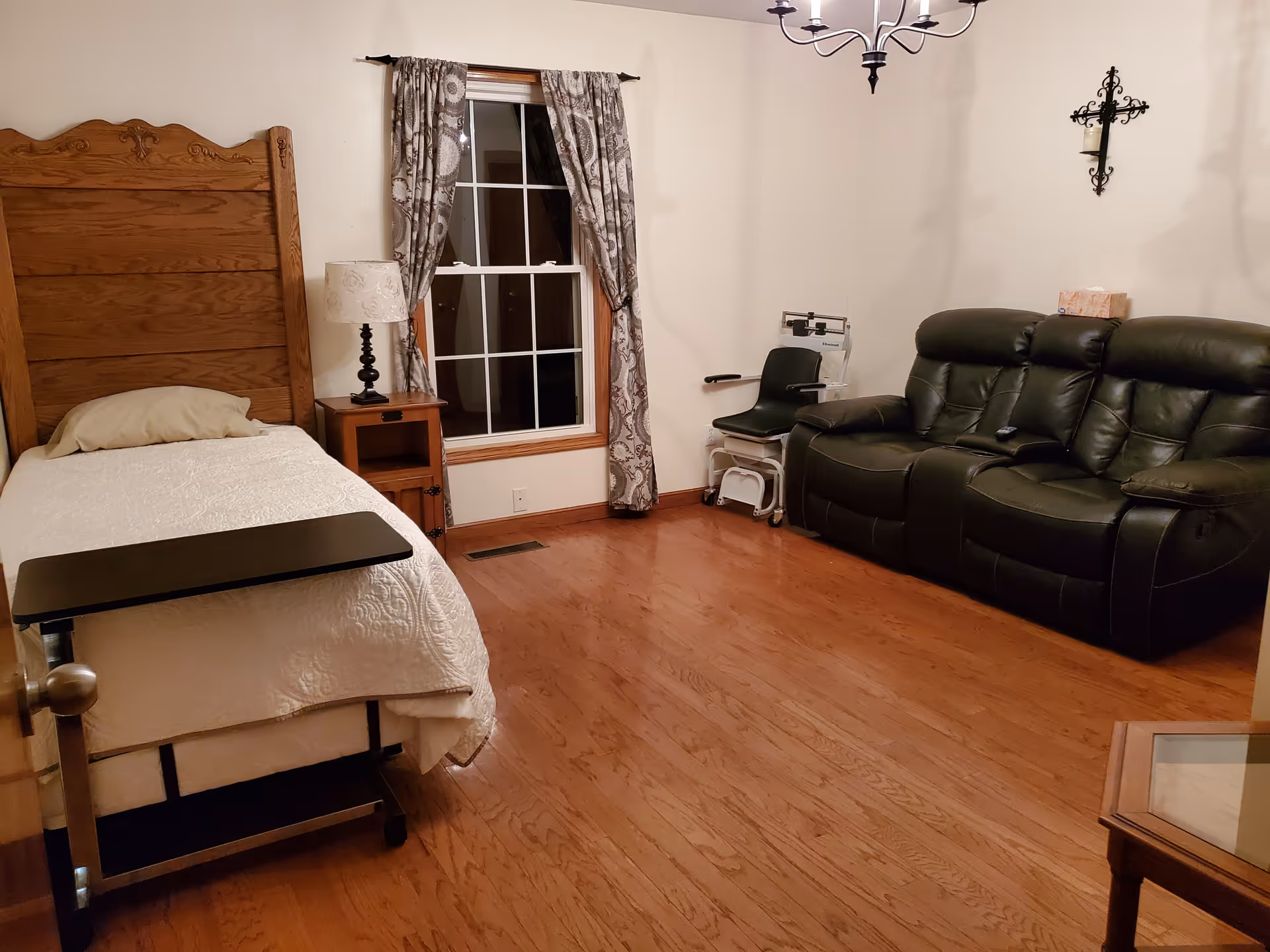 A bedroom with a single bed against a wooden headboard, nightstand and lamp, a window with patterned curtains, a leather reclining sofa, a medical transfer chair, hardwood floors and a wall cross.
