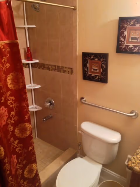 A bathroom with a white toilet, a beige tiled shower with a silver showerhead and faucet, a red and gold patterned shower curtain, a white corner shower caddy with a red bottle, a silver grab bar on the wall, and two framed decorative pictures hanging above the toilet.