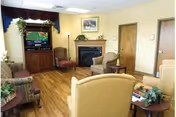 A cozy living room with wooden flooring, beige and patterned armchairs arranged around a fireplace. A TV is placed on a wooden cabinet in the corner, and there are plants and framed artwork on the walls. The room has a warm, inviting atmosphere with soft lighting.