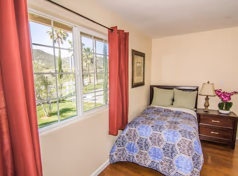 A small bedroom with a single bed covered in a blue and brown patterned quilt, two green pillows, a wooden nightstand with a lamp and a purple orchid, and a window with red curtains showing a view of palm trees and greenery outside.