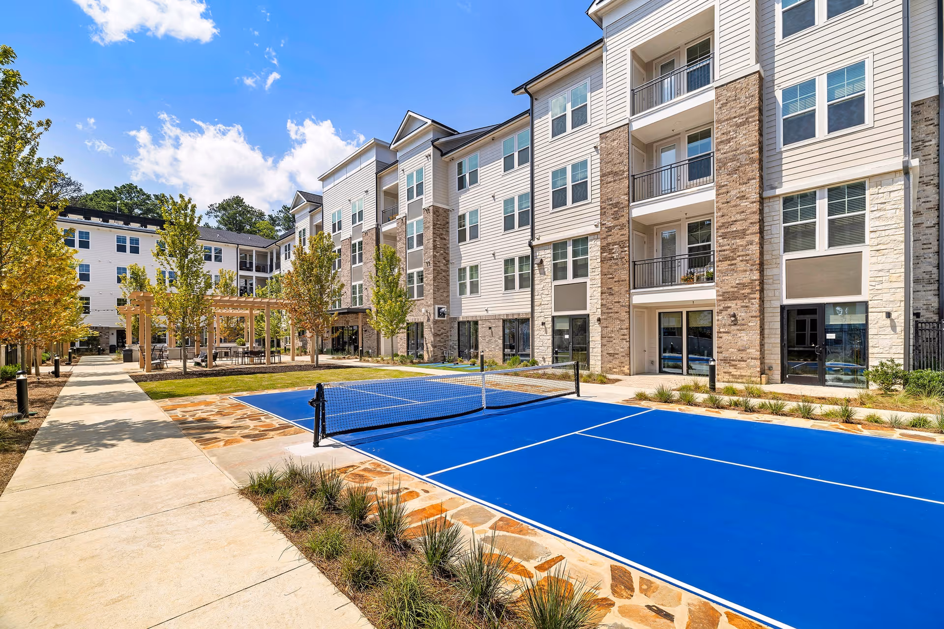 Outdoor view of a senior living facility courtyard with a bright blue pickleball court in the foreground, surrounded by a stone border and landscaping. The multi-story building with balconies and large windows is visible in the background under a clear blue sky with some clouds. There are trees and a pergola with seating along a paved walkway.