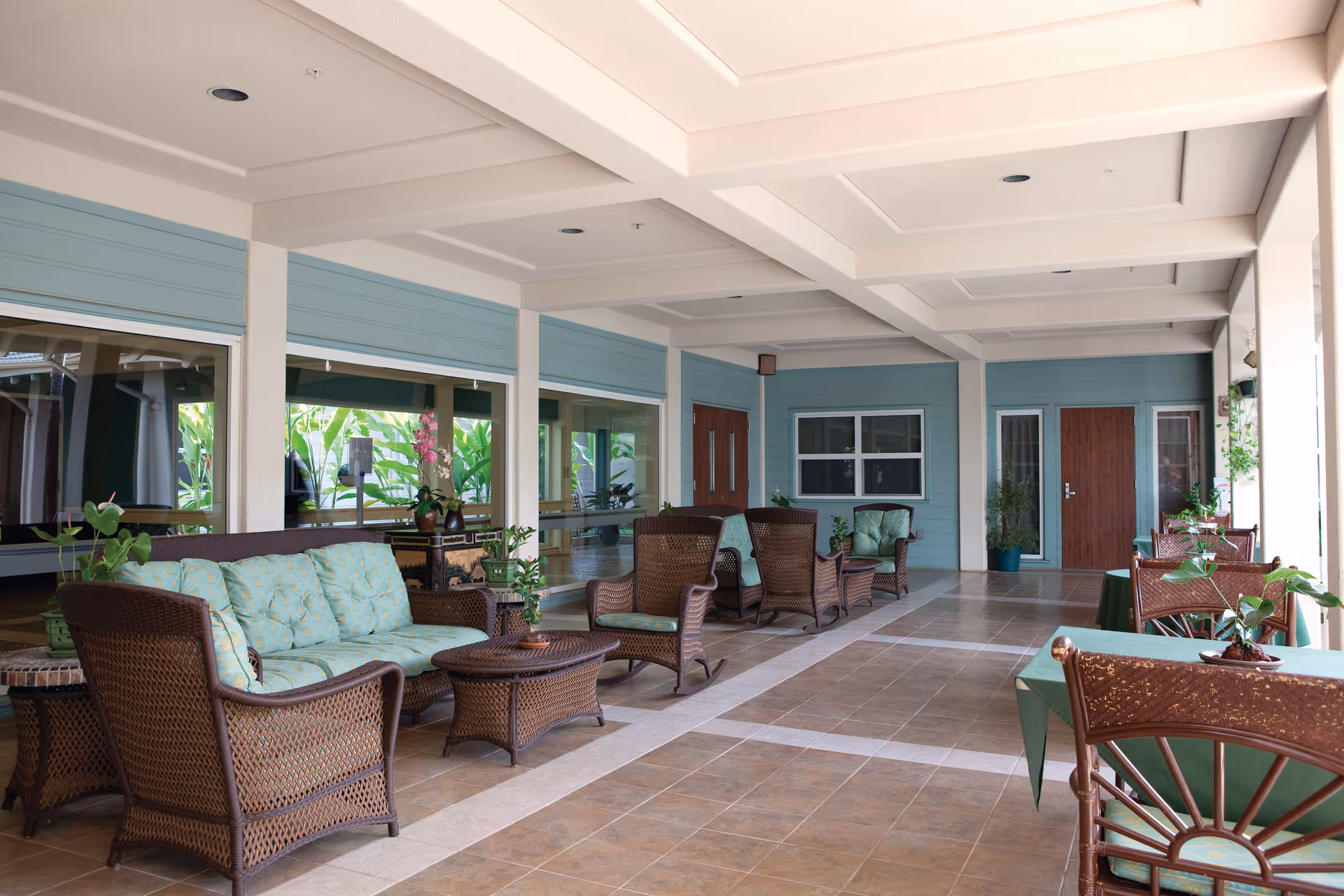 Covered outdoor seating area with wicker furniture including sofas and chairs with cushions, small tables, and potted plants. The area has tiled flooring, light blue walls, and wooden doors, with large windows reflecting greenery outside.
