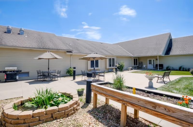 Outdoor patio area at a senior living facility with tables and chairs under umbrellas, a small raised garden bed with plants, a stone-bordered water feature with lily pads, a bench, and a barbecue grill against the building. The building has beige siding and a gray roof under a blue sky with some clouds.