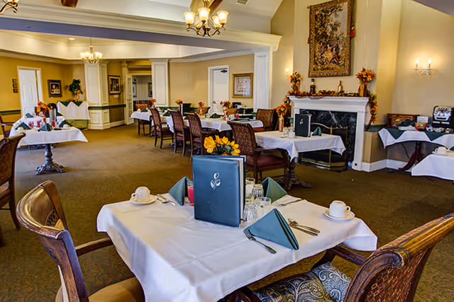 A dining room in a senior living facility with multiple tables covered in white tablecloths, set with cups, glasses, silverware, and green folded napkins. The room features wooden chairs, a fireplace decorated with autumn-themed garlands, a tapestry above the fireplace, and warm lighting from chandeliers and wall sconces.