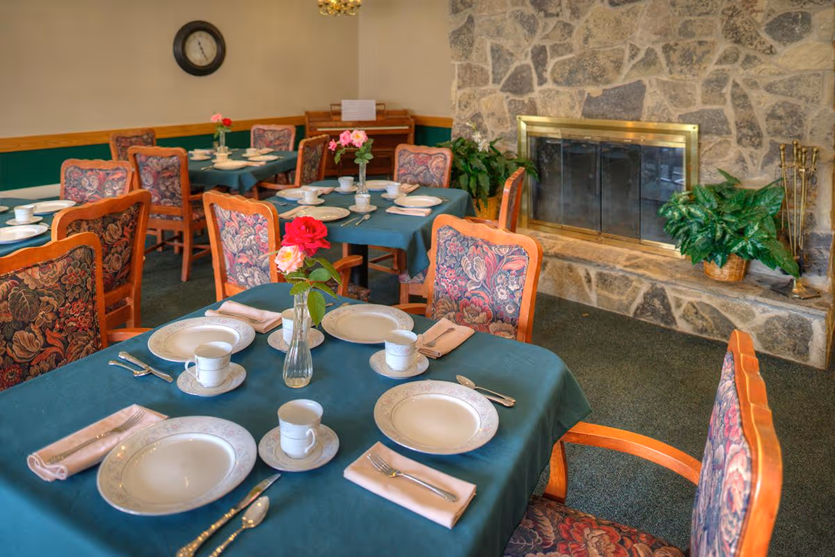 Dining room in a senior facility with tables set with plates and cups, floral-upholstered chairs, and a stone fireplace.
