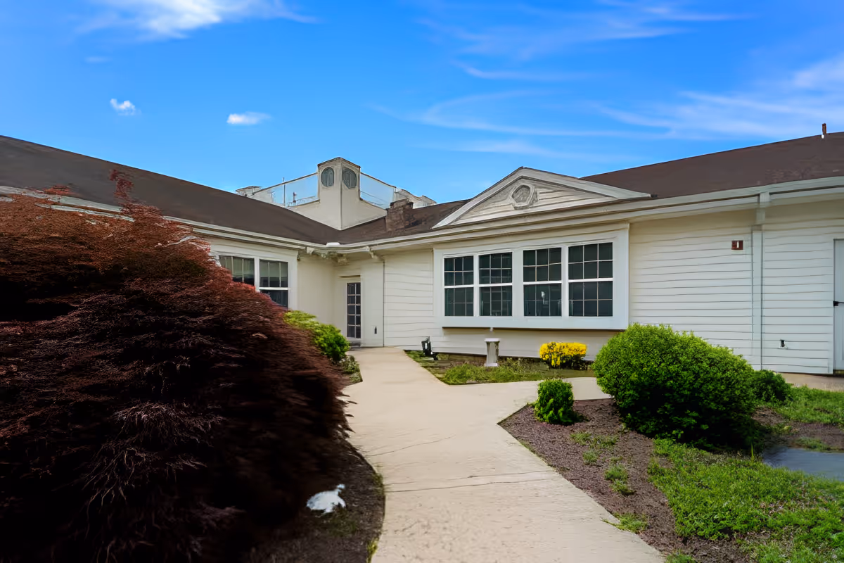 Exterior view of a single-story white building with a walkway, landscaped shrubs, and large windows under a blue sky.