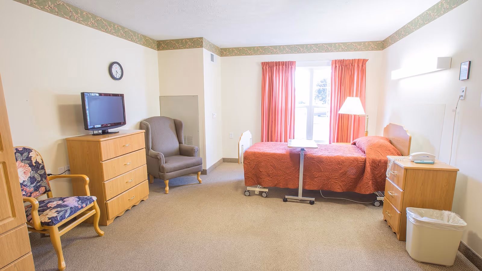 Well-lit senior bedroom with a single bed covered in a red bedspread, a dresser with a TV, chairs, and a window with red curtains.