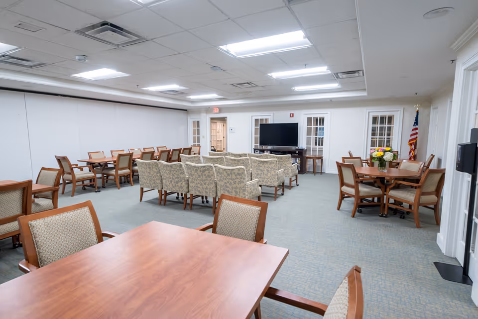 Spacious senior common room with multiple wooden dining tables, upholstered chairs, a seating area facing a TV, and an American flag.